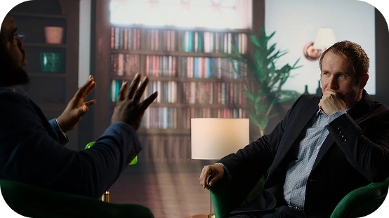 Twee mannen in pak voeren een serieus gesprek in een sfeervolle kamer met een boekenkast op de achtergrond.